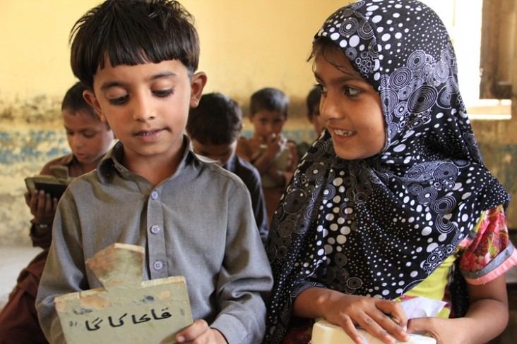 A young boy and a girl sit next to each other in a classroom. The boy is holding a small chalkboard and he is wearing a shirt